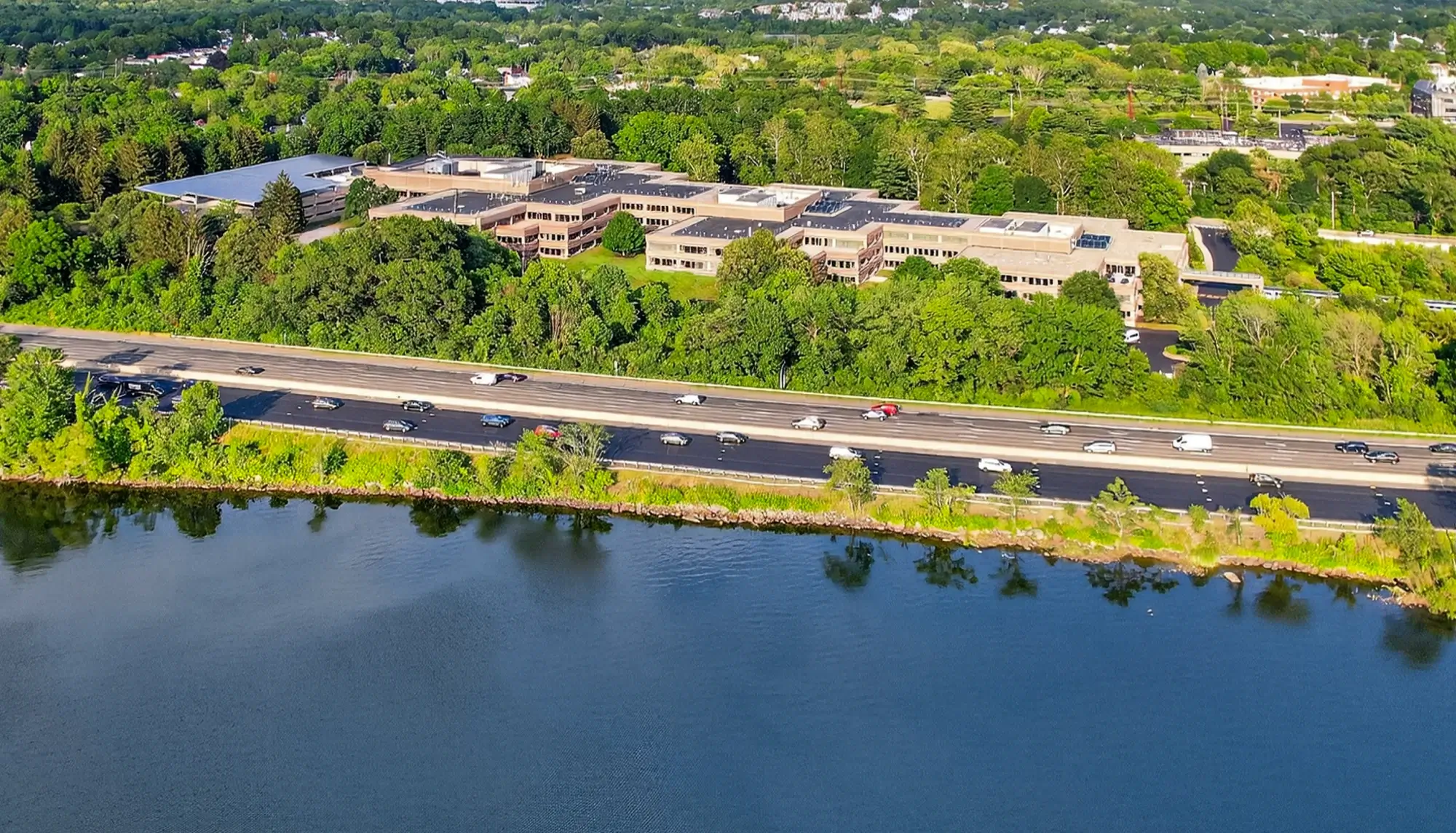 Elevated view of suburban campus bordered by forest, highway traffic, and reflective water surface